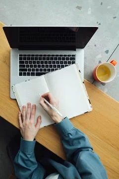 A woman takes notes in a notepad using a laptop sitting at the table, top view. Stock Photos