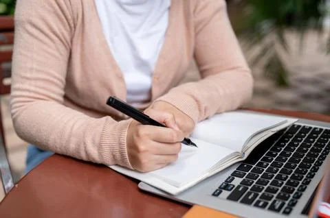 A woman taking notes in her notebook while studying online on her laptop at.. Stock Photos