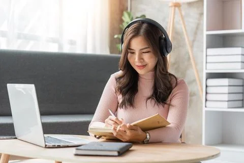 Woman taking notes in notebook while using laptop at home in living room 스톡 사진