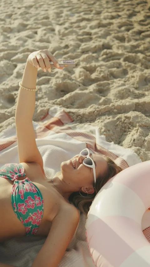 Woman taking a selfie while lying on a beach towel with a pool float nearby duri Stock Footage 339661406