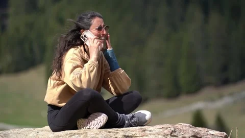 Woman talking on phone while sitting on rock in mountain landscape 스톡 동영상 307177882