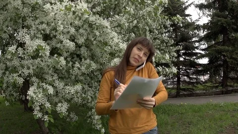 Woman talking on the phone working with documents next to blooming apple trees. Stock Footage 76794564