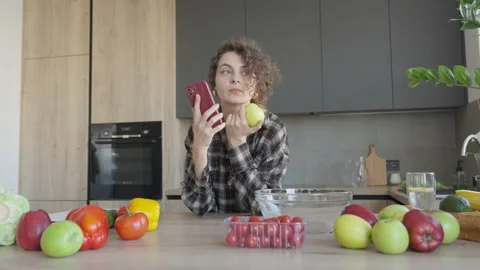 Woman Talking On Smartphone While Holding Fresh Apple In Kitchen Stock Footage 344698065