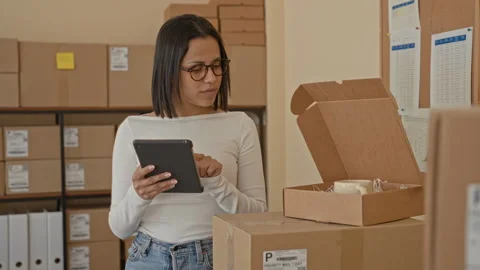 Woman tapping tablet while holding an open parcel box amid stacked shipping.. Stock Footage 337410266