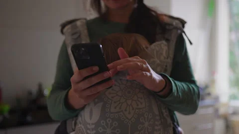 Woman taps smartphone screen while child rests in front carrier in home kit.. Stock-Footage 343662233