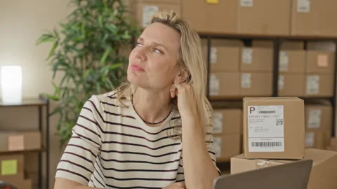 Woman touching ear beside stacked shipping parcels and laptop among labeled.. 動画素材 339254553