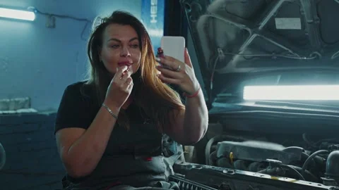 Woman touching up makeup while sitting on a car engine in garage Stock Footage 305848198