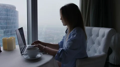 Woman types on notebook computer. Rainy cityscape view from skyscraper window Видео 97493015