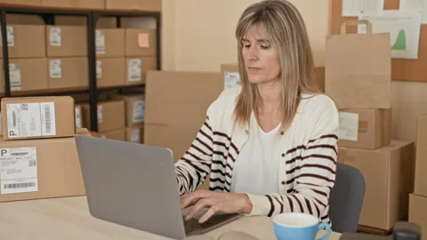 Woman typing on laptop while surrounded by stacked shipping parcels and pri.. Stock Footage 337432821