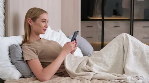 Woman uses smartphone while lying on bed in the morning at home. Stock Footage 253723359