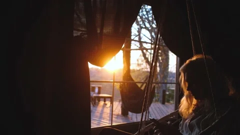 A woman uses a smartphone while sitting in a swing by the window at sunset Stock Footage 200820151