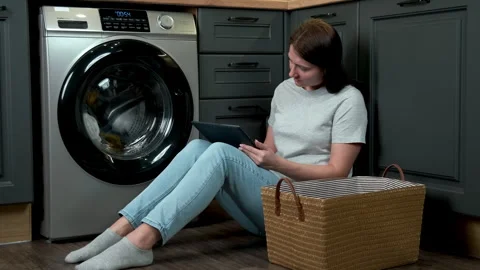 A woman uses a tablet while doing laundry while sitting next to a washing Stock Footage 301661055
