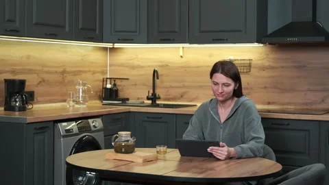 A woman uses a tablet while sitting at a table in the kitchen. Stock Footage 301661132