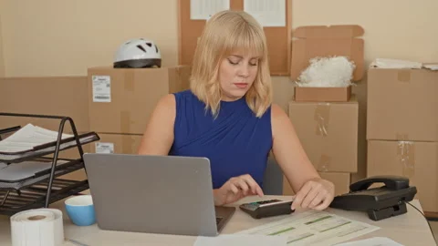 Woman using calculator with hand on keys beside laptop and stacked cardboar.. Stock Footage 337387834
