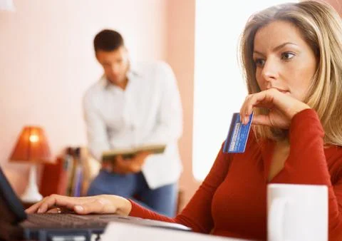 Woman using computer and holding credit card, man reading in background. Stock Photos