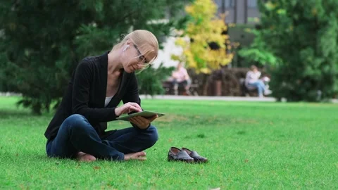 Woman using digital tablet sitting on green lawn in a city park. Stockbeeldmateriaal 215783589