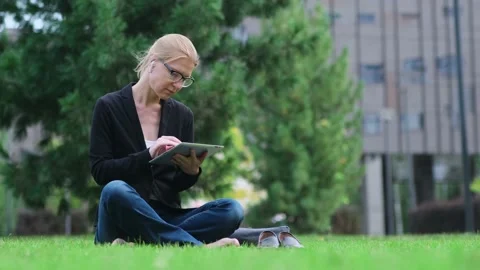 Woman using digital tablet sitting on green lawn in a city park. Stock Footage 220340516