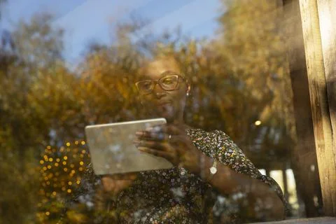 Woman using digital tablet at window with reflection of autumn trees 스톡 사진