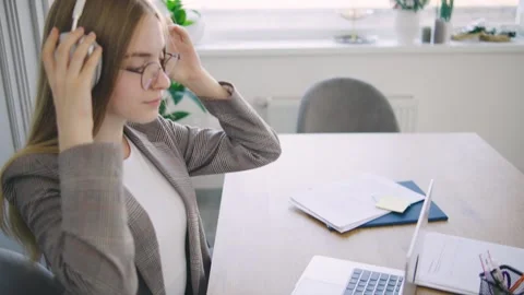 Woman using headphones while working at her desk with a laptop and notebooks in Stock Footage 302653221