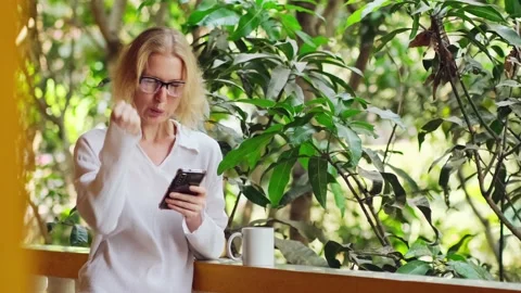 Woman is using her smartphone, drinking morning coffee on the balcony. Vidéo 236199719