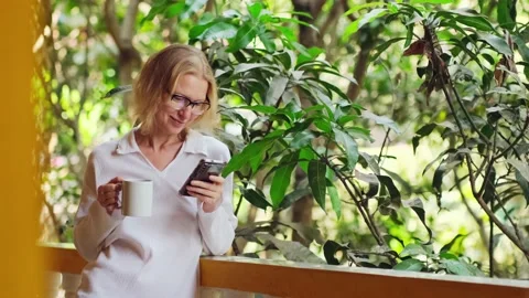 Woman is using her smartphone, drinking morning coffee on the balcony. Stockbeeldmateriaal 236816825
