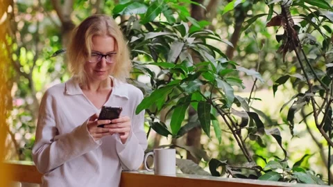 Woman is using her smartphone, drinking morning coffee on the balcony. Stockbeeldmateriaal 238508757