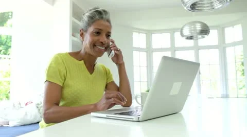 Woman Using Laptop And Talking On Phone In Kitchen At Home Stock Footage