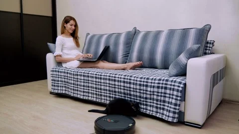 The woman using a laptop pc while a robotic vacuum cleaner is cleaning floor. 4K Stock-Footage 78172073