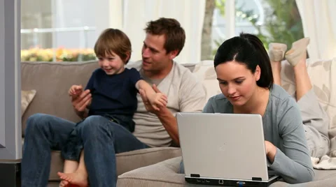 Woman using laptop while her boy and husband are watching tv 스톡 동영상 1018958