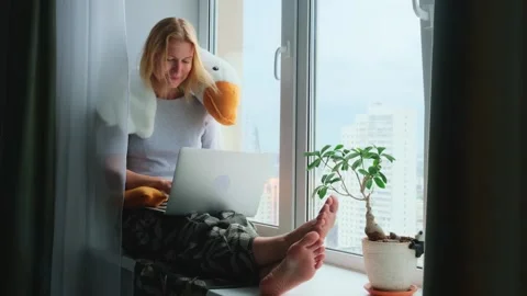 Woman using a laptop while sitting on a windowsill with a funny plush toy. Stock Footage 246334133