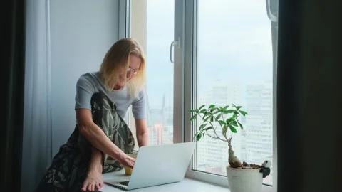 Woman using a laptop while sitting on a windowsill with a city view. Stockbeeldmateriaal 247381308