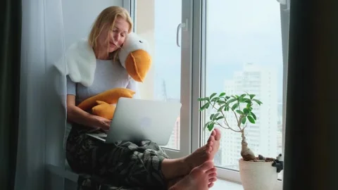 Woman using a laptop while sitting on a windowsill with a funny plush toy. Stock Footage 249640246
