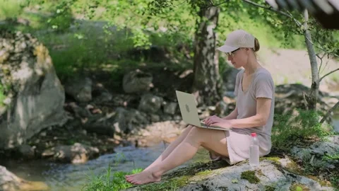 Woman using a laptop while sitting on the stone near mountain river. Stock Footage 313422201