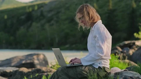 Woman using a laptop while sitting on the stone near mountain river. Stock Footage 314242784