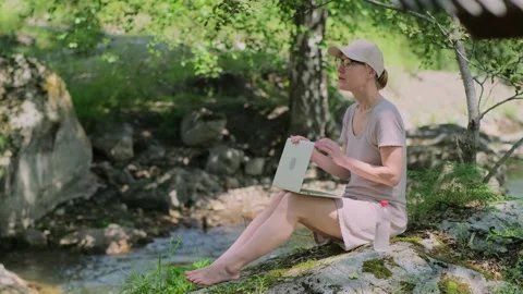 Woman using a laptop while sitting on the stone near mountain river. Stock Footage 315719834