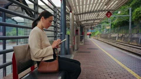 Woman using a mobile phone while sitting on a bench at a train station. 動画素材 338343241