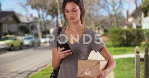 Woman using mobile phone while holding mail package on residential sidewalk Stock Photos