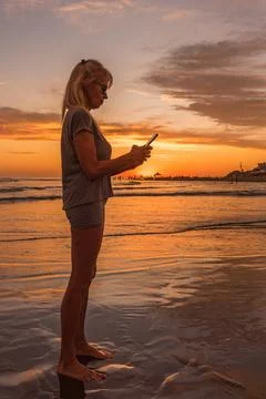 Woman using mobile phone while standing on the seashore at sunset. Stock Photos