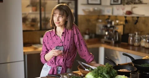 Woman using phone while standing with healthy food in the kitchen at home Stock Footage 173364190