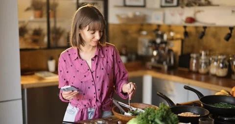 Woman using phone while standing with healthy food in the kitchen at home Stock Footage 208837525