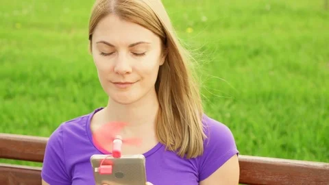 Woman using portable ventilator on mobile phone in park during heat. Cooling Stock Footage 77101337