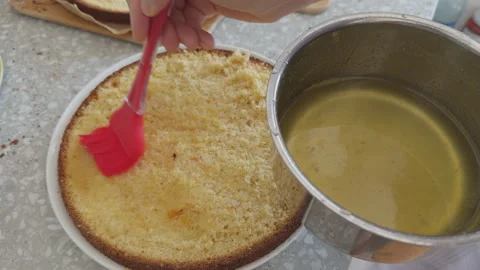 Woman using a red silicone brush to soak a fluffy sponge cake layer with syrup. Stock Footage 330434037