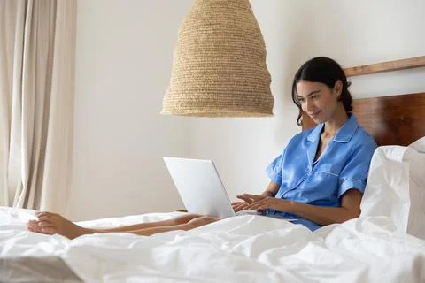 Woman using silver laptop while sitting on bed under woven pendant lamp in Foto stock