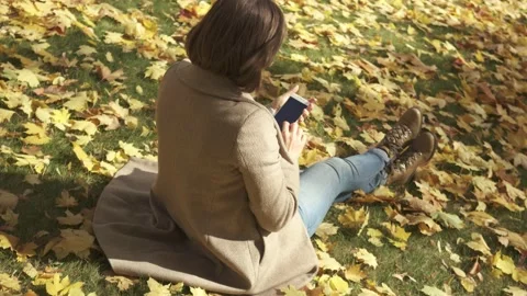 Woman using smart phone while sitting on grass in autumn park during sunny day Stock Footage 132285439