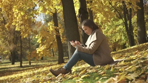 Woman using smart phone while sitting on the hill in autumn park Stock Footage 132286202