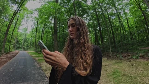 Woman using a smartphone on a forest path, symbolizing... 스톡 동영상 339101285