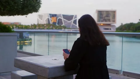 Woman using smartphone on waterfront deck overlooking modern theater building Video stock 335782160