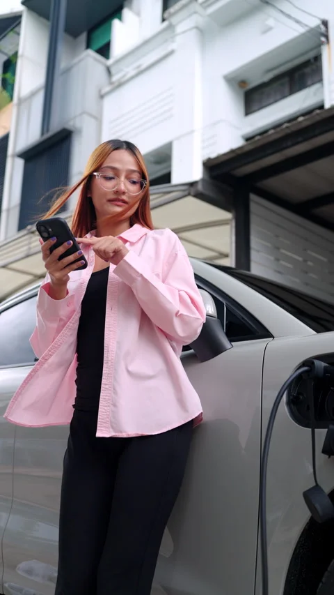 Woman using smartphone while her electric car is charging at home. Vídeos de archivo 346871803