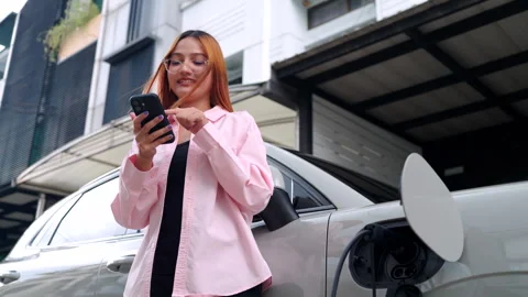 Woman using smartphone while her electric car is charging at home. 스톡 동영상 346871839