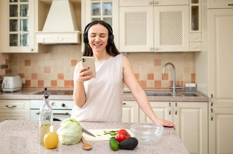 Woman using smartphone while preparing vegetables in kitchen. Wearing headpho Stock Photos
